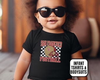 May include: A black short-sleeved infant bodysuit featuring a graphic of a gold football helmet with the letters SF, set against a black and white checkered square. The text SAN FRANCISCO FOOTBALL is printed in red. A white label reads INFANT TSHIRTS & BODYSUITS.