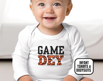 May include: A white long-sleeve infant bodysuit featuring the text GAME DEY printed on the chest. The word GAME is in solid black block letters, while the word DEY is in orange block letters with a black tiger stripe pattern throughout.