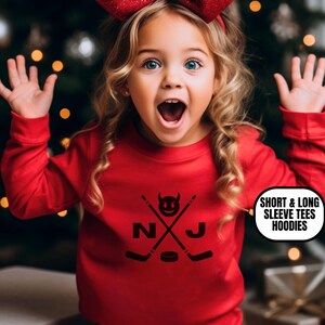 May include: A young child wearing a red long-sleeve shirt with a black graphic of crossed hockey sticks, a puck, and the letters "NJ". The child is wearing a large red bow and has their mouth open. Text on the image says "SHORT & LONG SLEEVE TEES HOODIES".