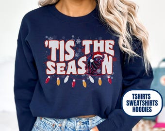 May include: Navy blue sweatshirt with the text "'Tis the Season" in white and red, featuring a buffalo wearing a Santa hat. The design includes snowflakes and string lights. The word "NAVY" is at the top left.