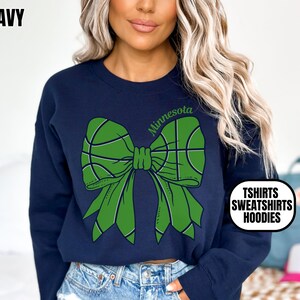 May include: Navy blue sweatshirt with a green bow design featuring basketballs. The word "Minnesota" is arched above the bow. The text "TSHIRTS SWEATSHIRTS HOODIES" is in a white circle. The word "NAVY" is in the upper left corner.
