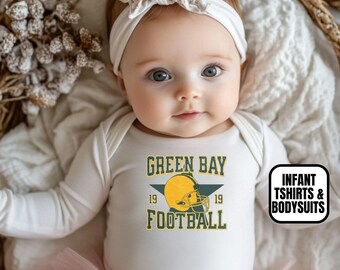 May include: A white long-sleeved bodysuit featuring a graphic of a yellow football helmet with the text GREEN BAY FOOTBALL 19 19. The garment is paired with a white floral headband and a light pink tulle skirt, creating a complete outfit ensemble.