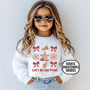 May include: A white long-sleeve sweatshirt featuring a graphic print with red bows, pom-poms, a football, a megaphone, and a foam finger. The text "Let's Go San Fran!" is printed in red below the graphics. The sweatshirt is paired with blue denim jeans.