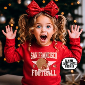 May include: A red long-sleeve sweatshirt featuring a white star graphic, a brown football helmet, and the text "SAN FRANCISCO 19 46 FOOTBALL". A small white oval label on the right side displays the black text "TSHIRTS SWEATSHIRTS HOODIES".