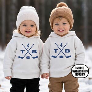 Pu&ograve; includere: Due bambini indossano felpe con cappuccio color crema con una stampa grafica blu raffigurante bastoni da hockey incrociati, un fulmine, un disco da hockey e le lettere T e B. Un bambino indossa un berretto crema, l'altro un berretto marrone.