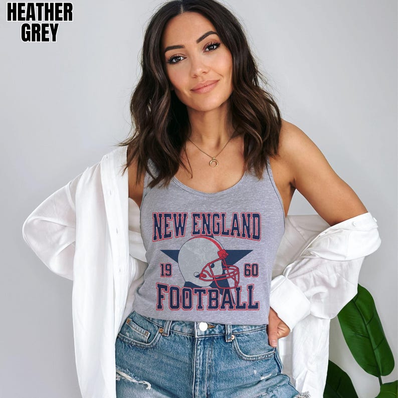 Pu&ograve; includere: Una canotta grigio melange con una stampa grafica che riporta il testo NEW ENGLAND FOOTBALL, i numeri 19 e 60 e l'illustrazione di un casco da football centrata su una stella blu scuro, indossata con una camicia bianca.