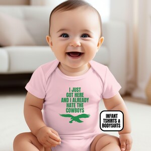 Puede incluir: Un body para beb&eacute; de color rosa claro con el texto verde I JUST GOT HERE AND I ALREADY HATE THE COWBOYS sobre un gr&aacute;fico de un &aacute;guila verde. La prenda tiene mangas cortas y cuello redondo, dise&ntilde;ada para la comodidad y el movimiento.