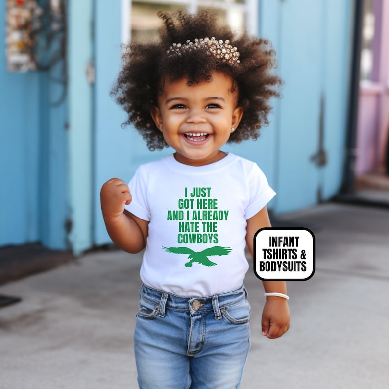 Puede incluir: Una camiseta blanca de manga corta con texto verde que dice I JUST GOT HERE AND I ALREADY HATE THE COWBOYS sobre un gr&aacute;fico de &aacute;guila verde. La camiseta se combina con jeans azules. Una etiqueta negra y blanca dice INFANT TSHIRTS & BODYSUITS.