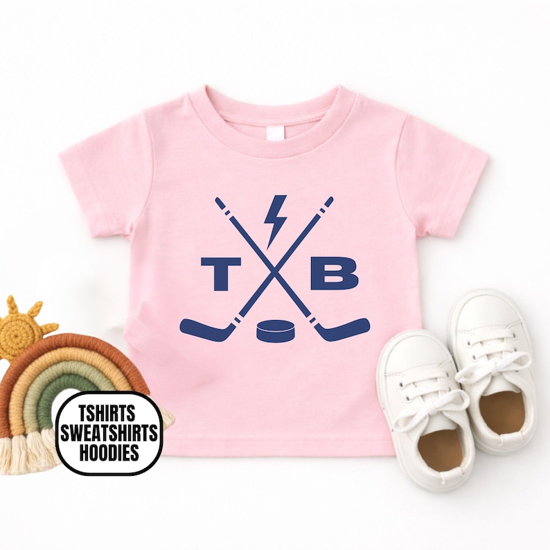 Pu&ograve; includere: Una t-shirt a maniche corte color rosa chiaro con una grafica blu scuro raffigurante due mazze da hockey incrociate, un fulmine, un disco da hockey e le lettere T e B. Il disegno &egrave; centrato sul petto della maglietta a girocollo.