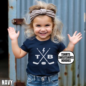 Pu&ograve; includere: Una t-shirt blu navy a maniche corte con una grafica bianca raffigurante due mazze da hockey incrociate, un fulmine, le lettere T e B e un disco da hockey. Il testo TSHIRTS SWEATSHIRTS HOODIES &egrave; stampato in un ovale bianco con bordo nero.
