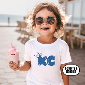 May include: A young child wearing sunglasses and a white t-shirt with a blue "KC" graphic and a crown above the letters. The child is holding a pink cupcake in a pink paper cone.