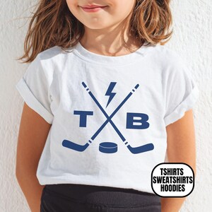 Pu&ograve; includere: Una t-shirt bianca a maniche corte con una grafica blu scuro di due mazze da hockey incrociate, un fulmine, un disco da hockey e le lettere T e B. La maglietta ha un girocollo, maniche arrotolate e un logo nell'angolo.