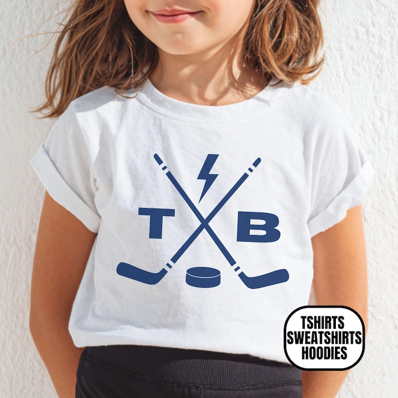Pu&ograve; includere: Una t-shirt bianca a maniche corte con una grafica blu scuro di due mazze da hockey incrociate, un fulmine, un disco da hockey e le lettere T e B. La maglietta ha un girocollo, maniche arrotolate e un logo nell'angolo.