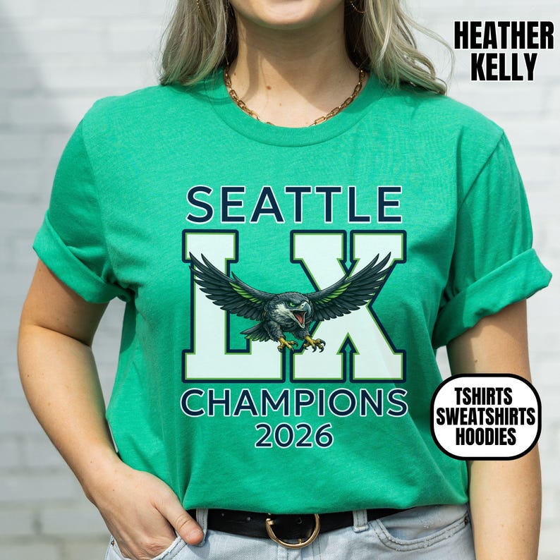 K&ouml;nnte beinhalten: Ein t&uuml;rkisgr&uuml;nes T-Shirt mit dem Text "SEATTLE" &uuml;ber einem stilisierten "LX"-Logo mit einer detaillierten Adlergrafik. Darunter steht "CHAMPIONS 2026". Das Shirt hat auch den Text "HEATHER KELLY" und eine Grafik mit der Aufschrift "TSHIRTS SWEATSHIRTS HOODIES".
