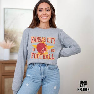 May include: A light grey heather long-sleeve sweatshirt with a vintage-style graphic of a football helmet and the text "Kansas City Football" with the numbers 19 and 60 on either side of the helmet.
