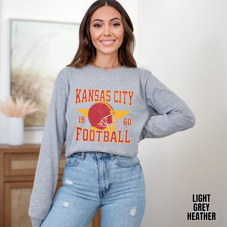 May include: A light grey heather long-sleeve sweatshirt with a vintage-style graphic of a football helmet and the text "Kansas City Football" with the numbers 19 and 60 on either side of the helmet.