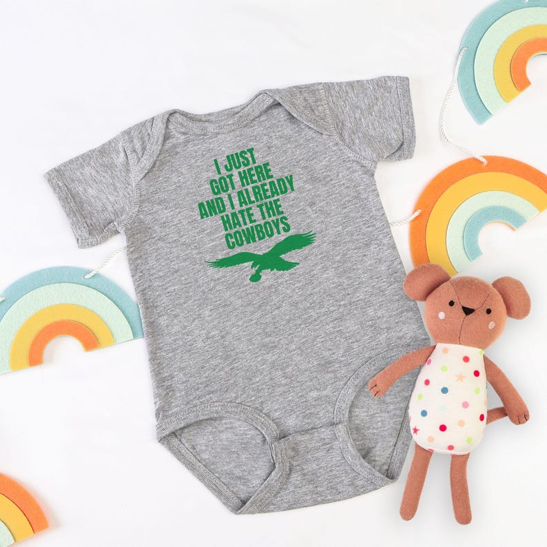 Puede incluir: Un body de beb&eacute; de manga corta en color gris jaspeado con el texto verde I JUST GOT HERE AND I ALREADY HATE THE COWBOYS sobre un gr&aacute;fico de &aacute;guila verde. La prenda tiene un cuello redondo y aberturas est&aacute;ndar para las piernas.