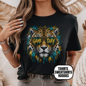 May include: A black short-sleeved t-shirt featuring a graphic of a leopard wearing sunglasses with the text GAME DAY printed on the lenses. The design includes blue and yellow paint splatter effects surrounding the leopard's face.