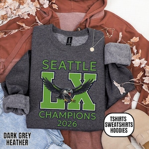 K&ouml;nnte beinhalten: Dunkelgraues meliertes Sweatshirt mit dem Text "SEATTLE LX CHAMPIONS 2026" in Gr&uuml;n und Wei&szlig;. Das "LX" zeigt eine Grafik eines Vogels mit ausgebreiteten Fl&uuml;geln. Das Sweatshirt hat auch den Text "TSHIRTS SWEATSHIRTS HOODIES".