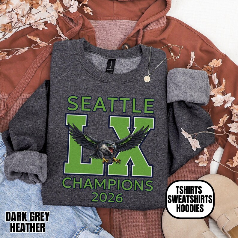 K&ouml;nnte beinhalten: Dunkelgraues meliertes Sweatshirt mit dem Text "SEATTLE LX CHAMPIONS 2026" in Gr&uuml;n und Wei&szlig;. Das "LX" zeigt eine Grafik eines Vogels mit ausgebreiteten Fl&uuml;geln. Das Sweatshirt hat auch den Text "TSHIRTS SWEATSHIRTS HOODIES".