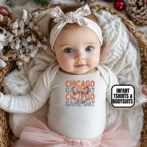May include: A baby wearing a white long-sleeved bodysuit with the text "CHICAGO GAME DAY" in orange and blue. The baby is also wearing a white headband with a bow and a pink tutu. The image includes a text box that says "INFANT TSHIRTS & BODYSUITS".