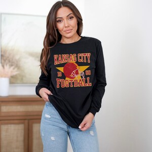 May include: Black long-sleeve shirt with a vintage-style graphic of a football helmet and the text "Kansas City Football" with the numbers 19 and 60 on either side of the helmet.