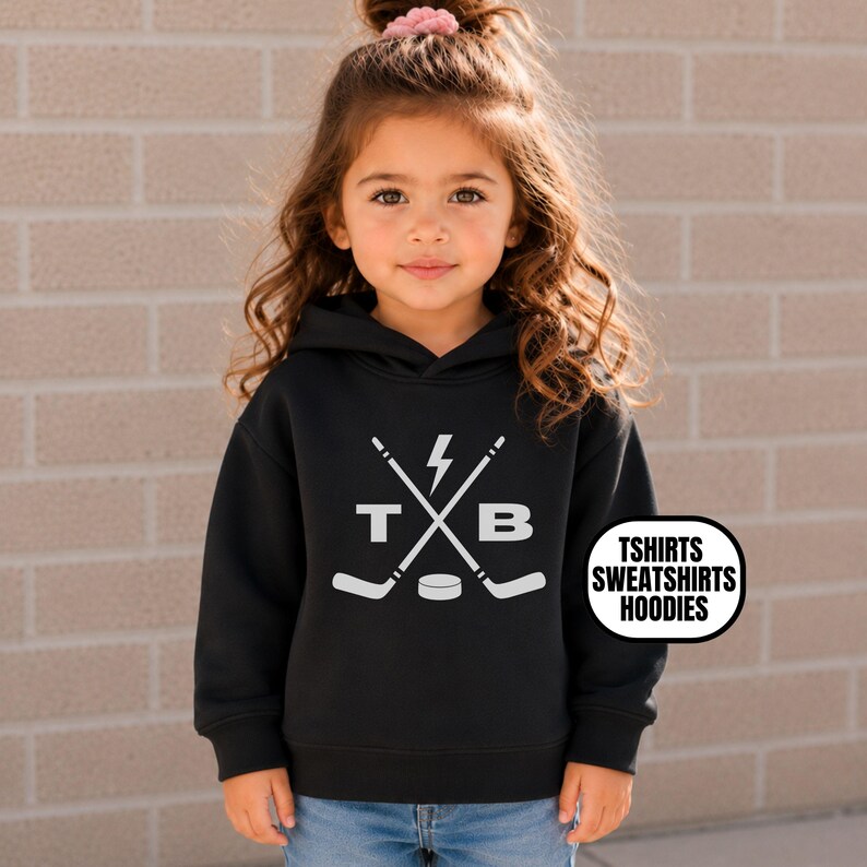 Pu&ograve; includere: Una felpa nera con cappuccio caratterizzata da una grafica bianca con due bastoni da hockey incrociati, un fulmine, un disco da hockey e le lettere T e B. Il testo TSHIRTS SWEATSHIRTS HOODIES &egrave; stampato in un ovale bianco sul lato destro.