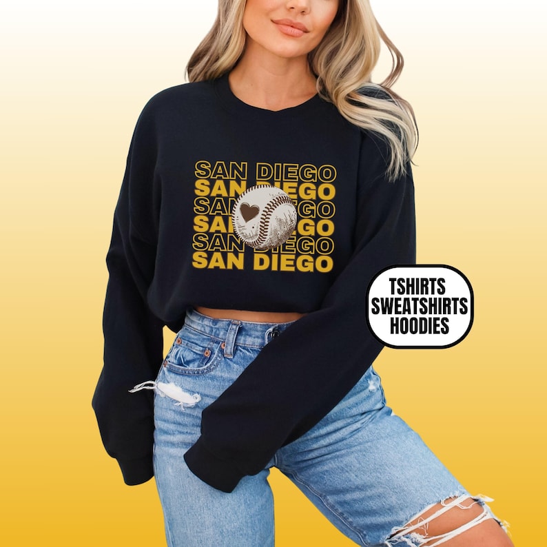 May include: A black cropped sweatshirt with a graphic of a baseball with a heart on it and the text "SAN DIEGO" repeated in yellow. The text "TSHIRTS SWEATSHIRTS HOODIES" is in a black box in the lower right corner.