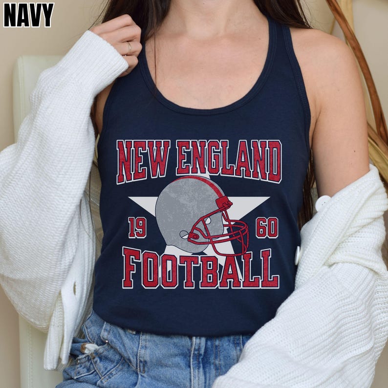 Pu&ograve; includere: Una canotta blu navy con una stampa grafica che riporta il testo NEW ENGLAND FOOTBALL in lettere rosse. Una stella bianca si trova dietro un casco da football americano grigio con maschera rossa. I numeri 19 e 60 sono stampati ai lati del casco.