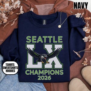 K&ouml;nnte beinhalten: Marineblaues Sweatshirt mit dem Text "SEATTLE LX CHAMPIONS 2026" in Gr&uuml;n und Wei&szlig;. Eine detaillierte Illustration eines Adlers mit ausgebreiteten Fl&uuml;geln ist mittig auf der Vorderseite. Das Sweatshirt hat auch den Text "TSHIRTS SWEATSHIRTS HOODIES".