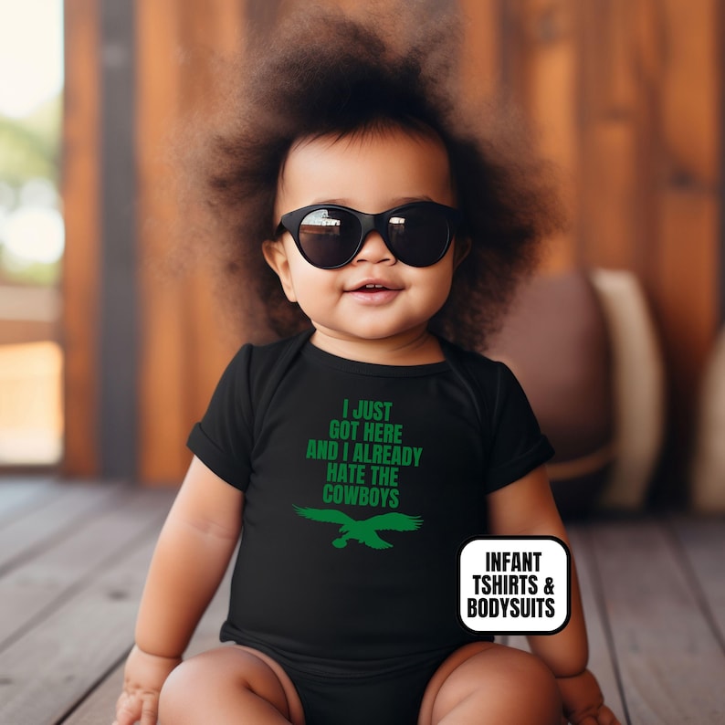 Puede incluir: Un body negro para beb&eacute; con texto verde que dice I JUST GOT HERE AND I ALREADY HATE THE COWBOYS sobre un gr&aacute;fico de &aacute;guila verde. La prenda incluye una etiqueta rectangular blanca con el texto INFANT TSHIRTS & BODYSUITS.