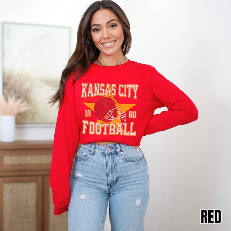 May include: A red long-sleeved cropped sweatshirt with the text 'Kansas City Football' and a football helmet graphic. The text '19' and '60' are on either side of the helmet graphic. The sweatshirt is worn by a person with long brown hair. The person is wearing blue jeans.