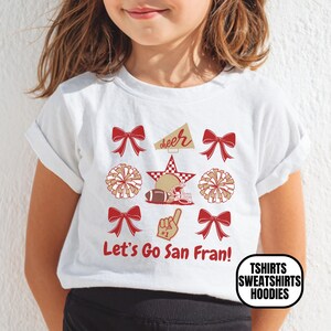 May include: A white short-sleeved t-shirt featuring a red and gold graphic print. The design includes four red bows, two pom-poms, a megaphone with the word cheer, a football helmet, a star, and a foam finger. The text Let's Go San Fran! is printed below.