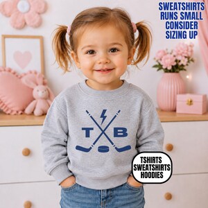Pu&ograve; includere: Una felpa girocollo grigio melange con una grafica blu scuro raffigurante due mazze da hockey incrociate, un fulmine, un disco da hockey e le lettere T e B. Il testo sull'immagine recita: SWEATSHIRTS RUNS SMALL CONSIDER SIZING UP e TSHIRTS SWEATSHIRTS HOODIES.