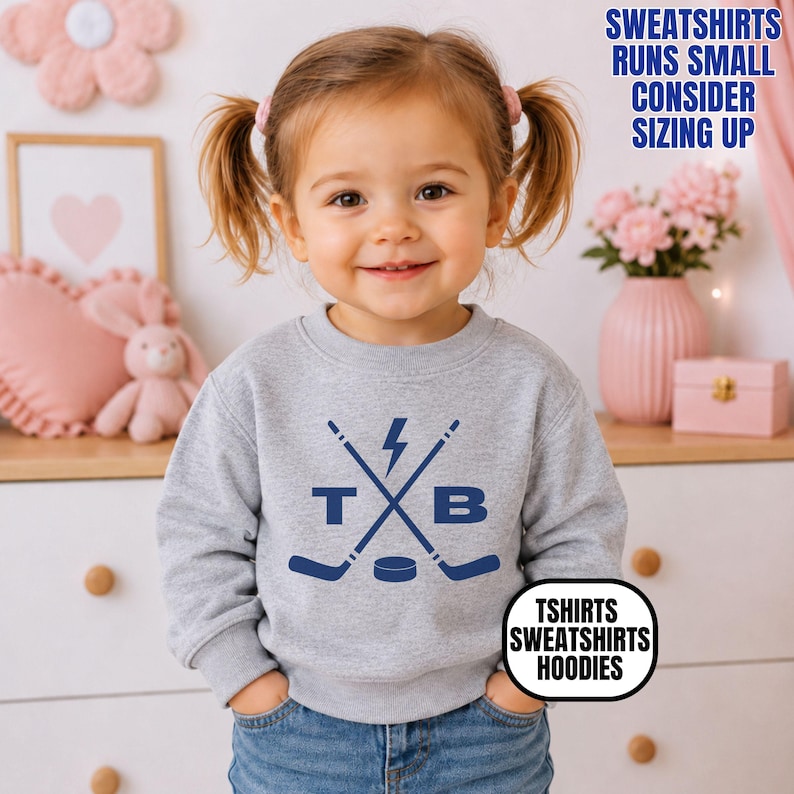 Pu&ograve; includere: Una felpa girocollo grigio melange con una grafica blu scuro raffigurante due mazze da hockey incrociate, un fulmine, un disco da hockey e le lettere T e B. Il testo sull'immagine recita: SWEATSHIRTS RUNS SMALL CONSIDER SIZING UP e TSHIRTS SWEATSHIRTS HOODIES.