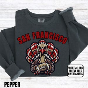 May include: Pepper-colored sweatshirt with the text "SAN FRANCISCO" in red. The graphic features a turkey holding a football. The sweatshirt has long sleeves and the text "SHORT & LONG SLEEVE TEES SWEATSHIRTS".