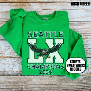 K&ouml;nnte beinhalten: Irisch gr&uuml;nes Sweatshirt mit dem Text "SEATTLE LX CHAMPIONS 2026" und einer Adlergrafik. Das Sweatshirt hat auch die Worte "TSHIRTS SWEATSHIRTS HOODIES" in einem wei&szlig;en Oval.