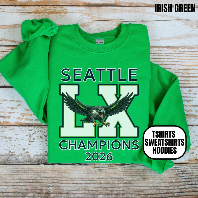 K&ouml;nnte beinhalten: Irisch gr&uuml;nes Sweatshirt mit dem Text "SEATTLE LX CHAMPIONS 2026" und einer Adlergrafik. Das Sweatshirt hat auch die Worte "TSHIRTS SWEATSHIRTS HOODIES" in einem wei&szlig;en Oval.