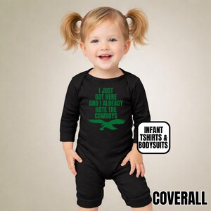 Puede incluir: Un body negro de manga larga para beb&eacute; con el texto verde I JUST GOT HERE AND I ALREADY HATE THE COWBOYS sobre un gr&aacute;fico de &aacute;guila verde. La prenda incluye una peque&ntilde;a etiqueta blanca con el texto INFANT TSHIRTS & BODYSUITS.