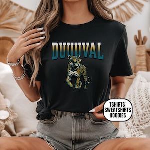May include: Black t-shirt featuring the word "DUUUVAL" in teal and gold lettering above a leopard wearing sunglasses. The leopard has a yellow and black spotted pattern. The shirt also has a circular logo that says "TSHIRTS SWEATSHIRTS HOODIES".