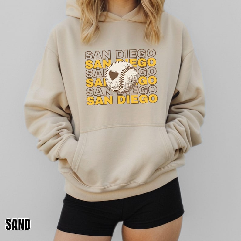 May include: A beige hoodie with a pocket and a baseball graphic with a heart on it. The text "SAN DIEGO" is repeated in yellow and black on the hoodie.