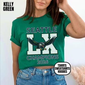 K&ouml;nnte beinhalten: Kelly-gr&uuml;nes T-Shirt mit dem Text "SEATTLE LX CHAMPIONS 2026" und einer Grafik eines Vogels mit ausgebreiteten Fl&uuml;geln. Das Shirt hat auch die Worte "TSHIRTS SWEATSHIRTS HOODIES" in einem wei&szlig;en Kreis.