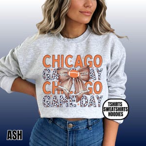 May include: Ash gray sweatshirt with the words "CHICAGO GAME DAY" in orange and leopard print. A football-shaped bow is centered on the design. The text "TSHIRTS SWEATSHIRTS HOODIES" is in a white circle.