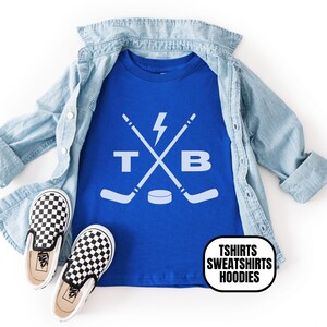 Pu&ograve; includere: Una t-shirt blu reale con una grafica bianca di due bastoni da hockey incrociati, un fulmine, un disco da hockey e le lettere T e B. La maglietta &egrave; sovrapposta a una camicia di jeans azzurra con un paio di scarpe slip-on a scacchi bianchi e neri.