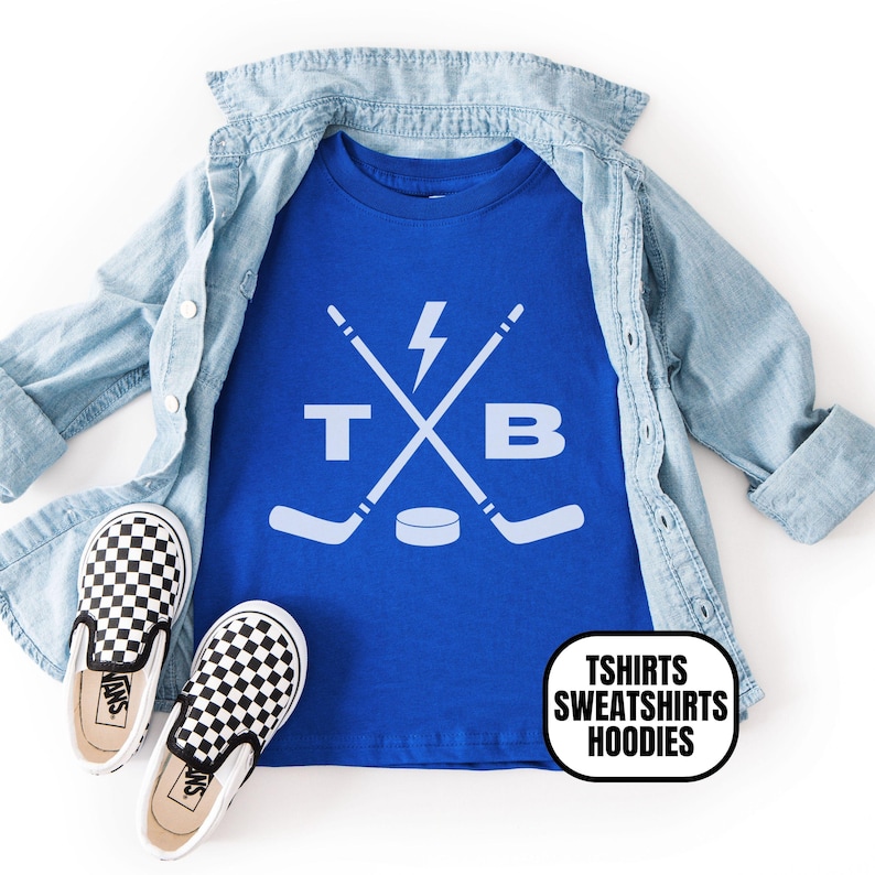Pu&ograve; includere: Una t-shirt blu reale con una grafica bianca di due bastoni da hockey incrociati, un fulmine, un disco da hockey e le lettere T e B. La maglietta &egrave; sovrapposta a una camicia di jeans azzurra con un paio di scarpe slip-on a scacchi bianchi e neri.