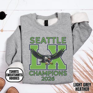 K&ouml;nnte beinhalten: Hellgraues Heather-Sweatshirt mit dem Text "SEATTLE LX CHAMPIONS 2026" in Gr&uuml;n und Blau. Das Design zeigt einen stilisierten Adler. Das Sweatshirt hat einen Rundhalsausschnitt und lange &Auml;rmel.