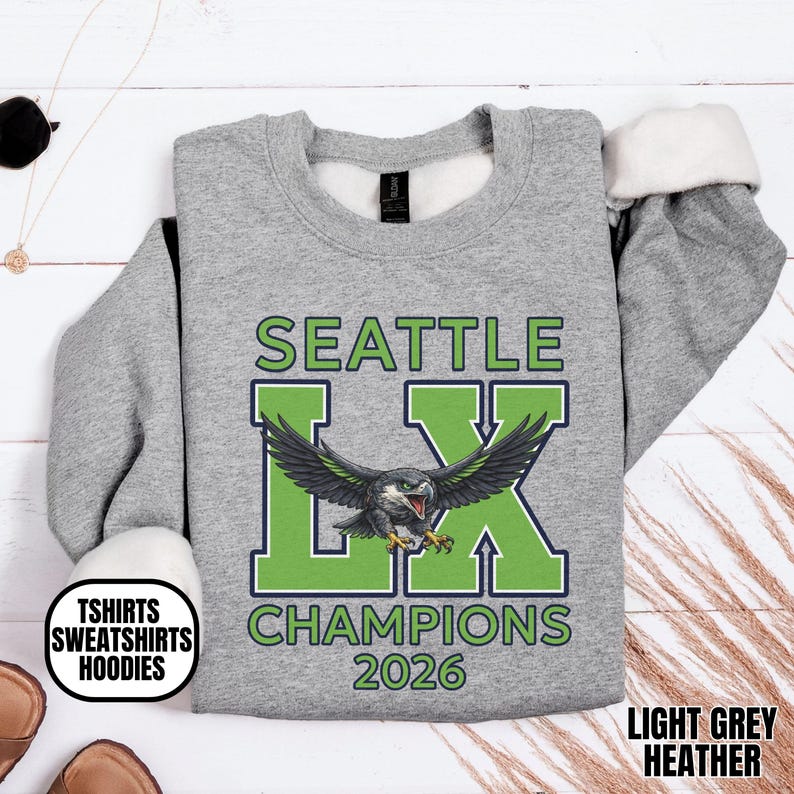 K&ouml;nnte beinhalten: Hellgraues Heather-Sweatshirt mit dem Text "SEATTLE LX CHAMPIONS 2026" in Gr&uuml;n und Blau. Das Design zeigt einen stilisierten Adler. Das Sweatshirt hat einen Rundhalsausschnitt und lange &Auml;rmel.
