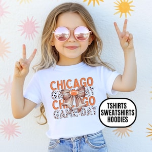 May include: A white short-sleeved t-shirt featuring the orange text CHICAGO GAME DAY with a brown leopard-print bow and a football graphic in the center. A black rounded rectangular label displays the white text TSHIRTS SWEATSHIRTS HOODIES.