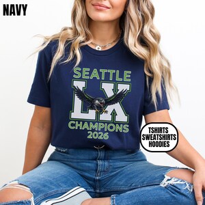K&ouml;nnte beinhalten: Marineblaues T-Shirt mit dem Text "SEATTLE LX CHAMPIONS 2026" und einer Vogelgrafik. Das Shirt hat auch einen wei&szlig;en Kreis mit dem Text "TSHIRTS SWEATSHIRTS HOODIES".