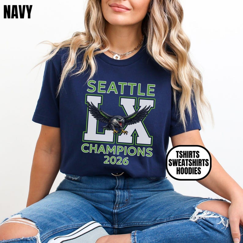 K&ouml;nnte beinhalten: Marineblaues T-Shirt mit dem Text "SEATTLE LX CHAMPIONS 2026" und einer Vogelgrafik. Das Shirt hat auch einen wei&szlig;en Kreis mit dem Text "TSHIRTS SWEATSHIRTS HOODIES".